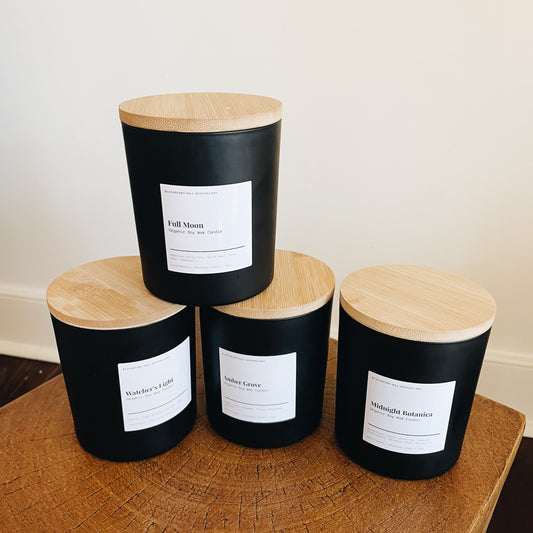 Four black soy-wax candles with wooden lids, arranged on a wooden surface, with white labels on each candle.