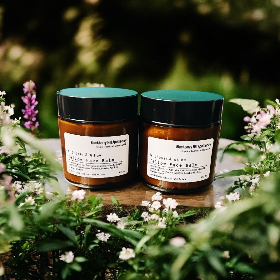 Two jars of 'Blackberry Hill Apothecary' Yellow Face Balm on a wooden surface with flowers in the background.