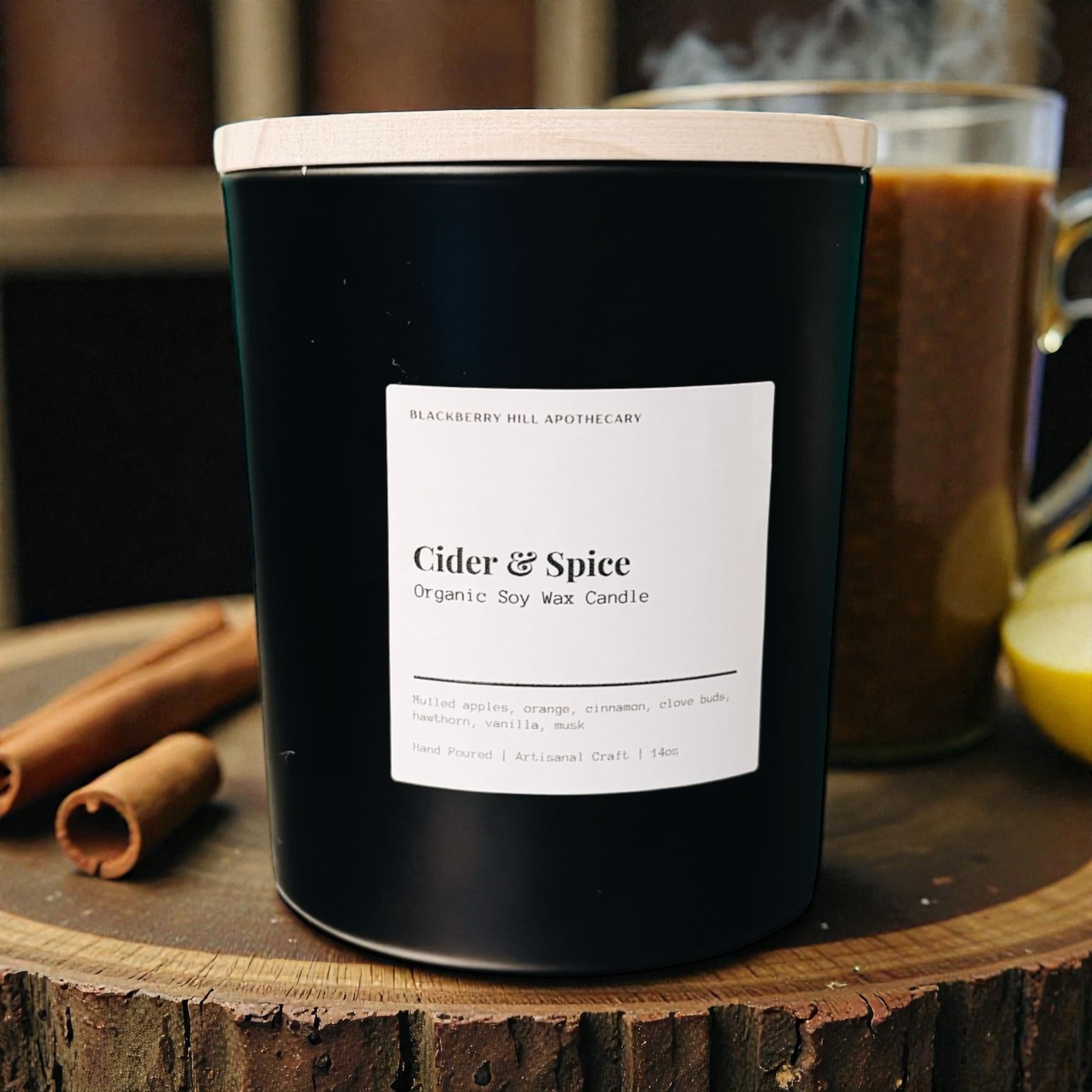 Black candle labeled 'Cider & Spice' on a wooden surface with cinnamon sticks and a mug in the background.